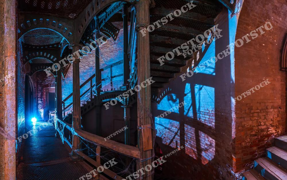 На фотографии показан интерьер промышленного здания с характерными металлическими конструкциями и кирпичными стенами. Пространство освещено таинственным синим светом, который подчеркивает детали архитектуры и создаёт игру теней на кирпичной кладке. Металлические лестницы и перекрытия напоминают эпоху промышленной революции, что добавляет снимку индустриального духа. Атмосфера фотографии навевает мысли о прошлом и многолетней истории здания.